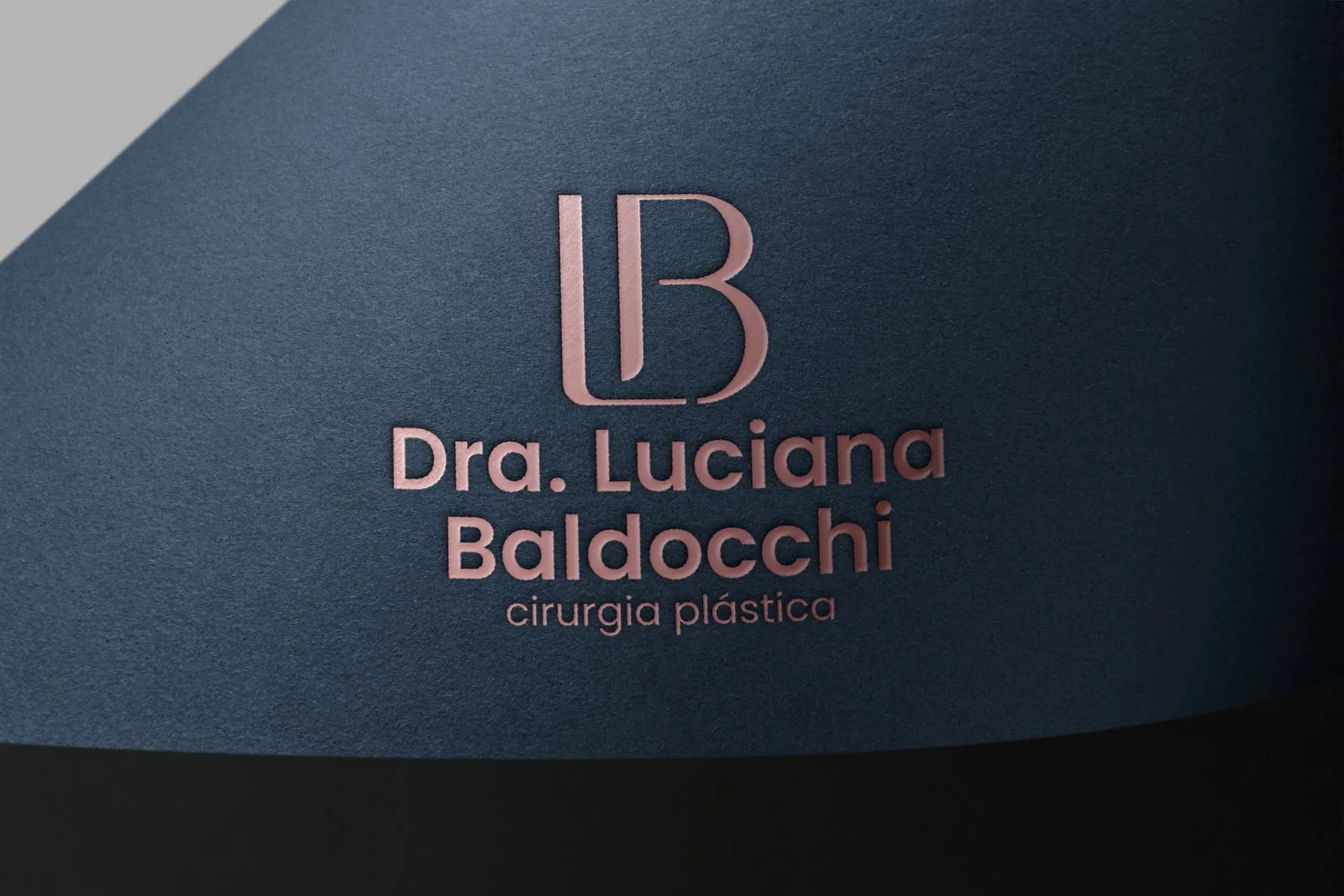 Monograma LB em hot stamping rosé sobre papel texturizado azul-marinho, com a assinatura Dra. Luciana Baldocchi
